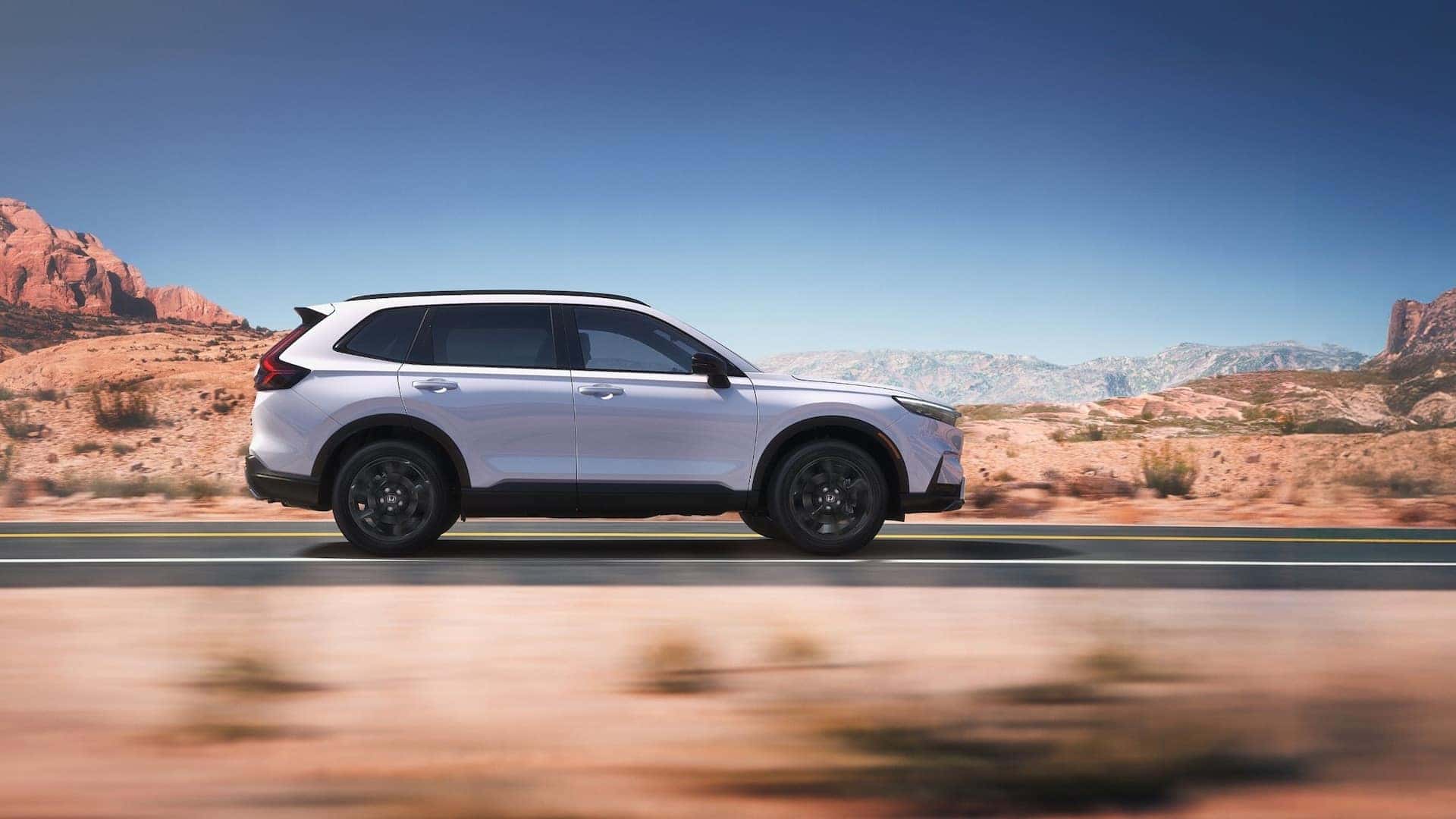 A silver Honda CR-V Hybrid in profile driving down the road in a desert.