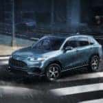 A light blue 2023 Honda HR-V Sport is shown driving through and intersection in the rain.