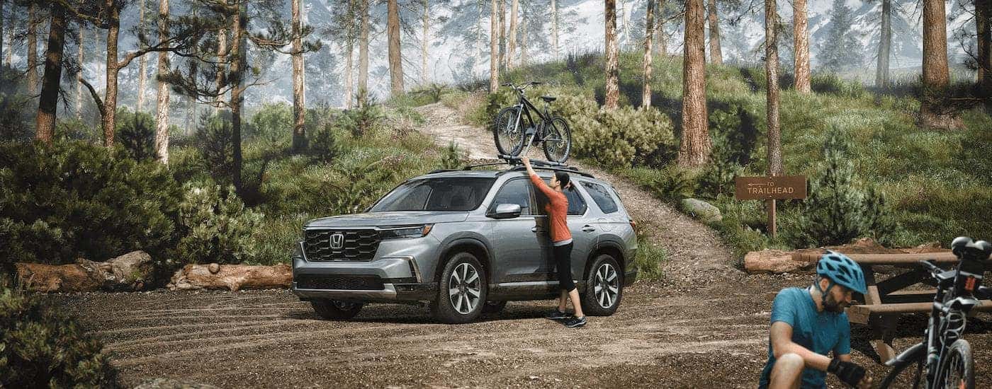 A woman is shown adjusting a bike on the roof rack of a grey 2023 Honda Pilot Trailsport.