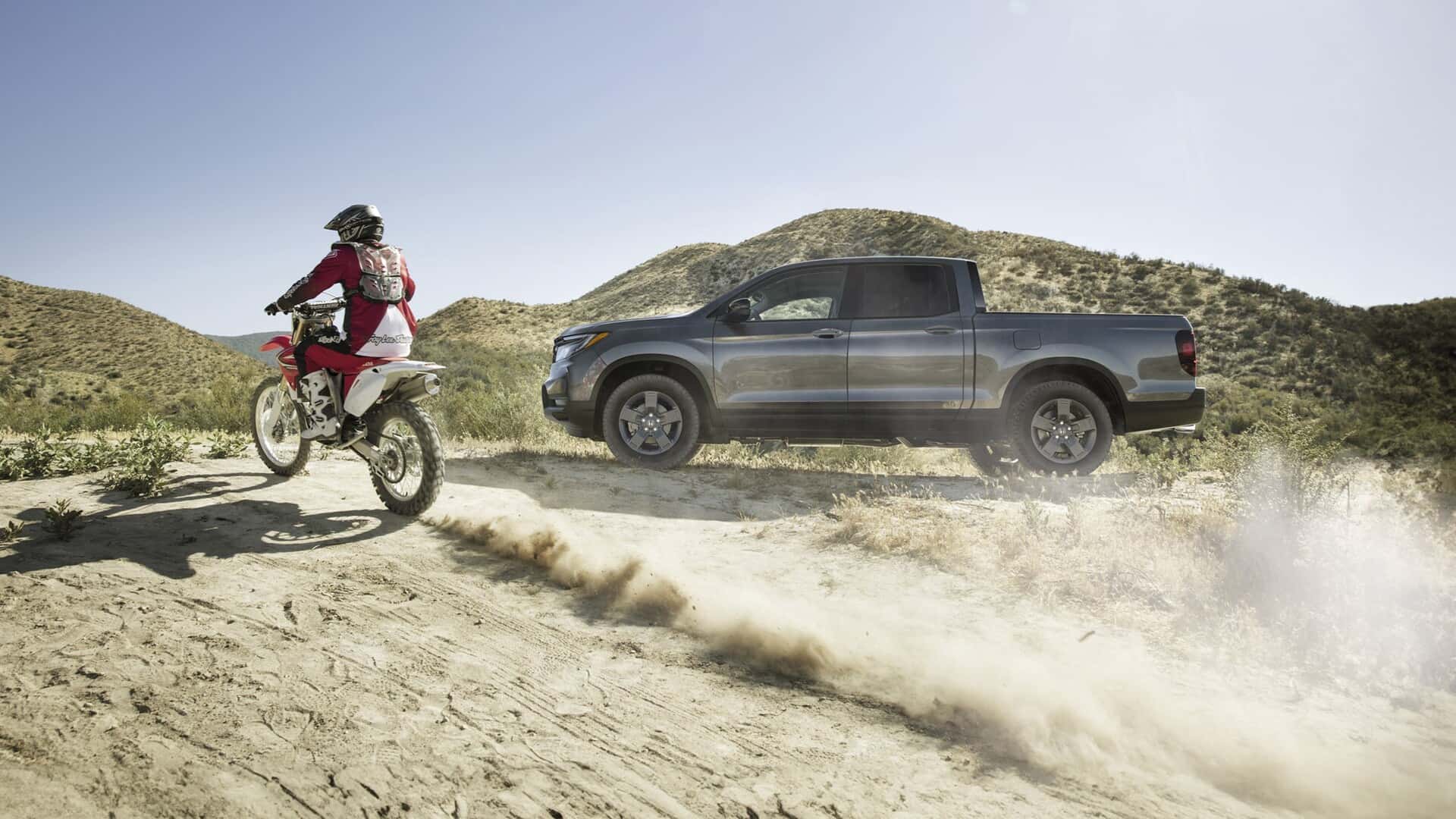 A gray Honda Ridgeline parked in a desert while a man on a dirt bike drives by