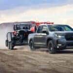 A gray Honda Ridgeline driving over a dirt road while towing a trailer with dirt bikes and golf carts