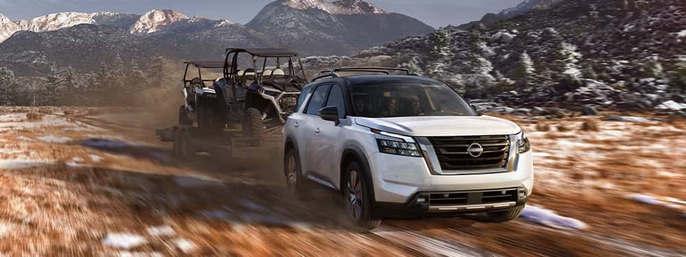 A white 2025 Nissan Pathfinder towing a flatbed with two carts through a snowy mountainous region