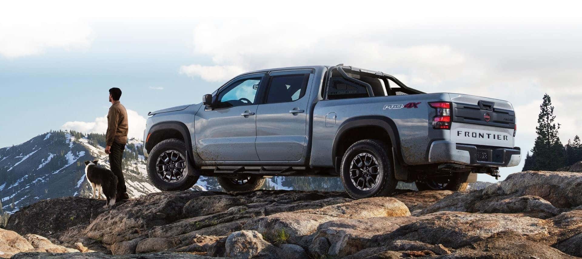 A silver Nissan Frontier at the top of a Rocky Mountain, with a man and his dog standing in front of it