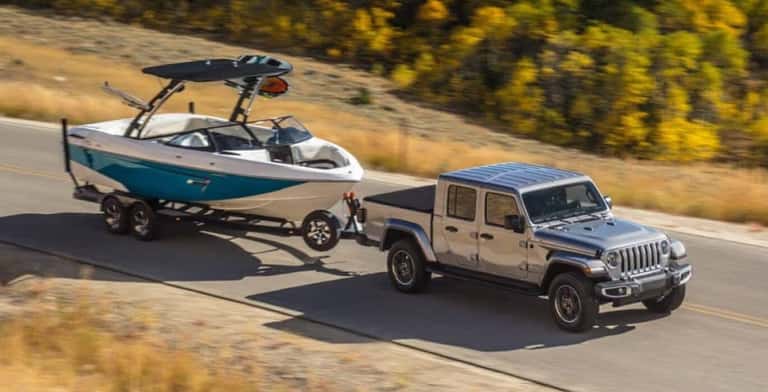 Wide shot of a silver Jeep Gladiator towing a large boat on a country road.