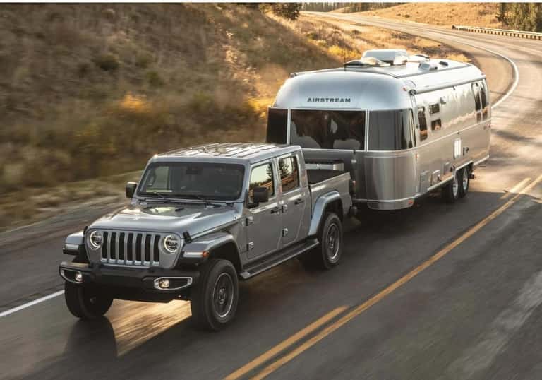 Front action shot of a silver Jeep Gladiator towing a camper trailer on a sunny highway.