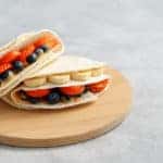 Two tortillas with different fillings of peanut butter, banana, strawberry, blueberry, almond on cutting board