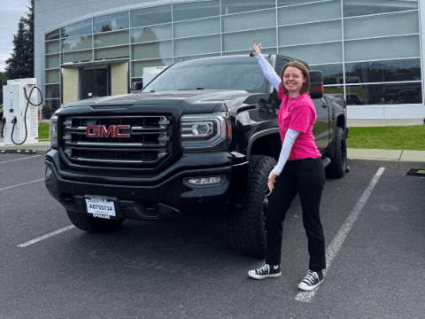 A girl with arms opened wide standing by a used black 2017 GMC Sierra truck parked on pavement