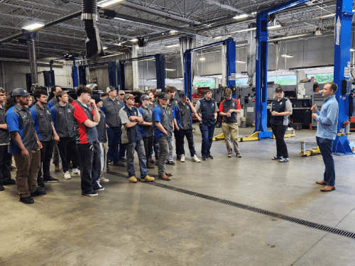 NEWTech Prep Auto Department students listening to a presentation in a professional auto repair bay with car lifts.
