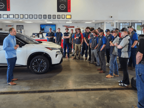 NEWTech Prep Auto Department students receiving training next to a new white Nissan SUV in the dealership showroom.