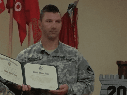 A soldier in a US Army uniform holding a certificate during a military ceremony.