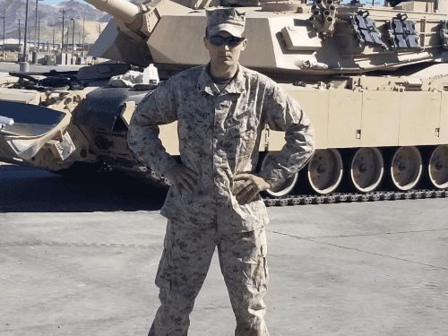 A soldier wearing desert camouflage uniform, standing in front of a large tank.