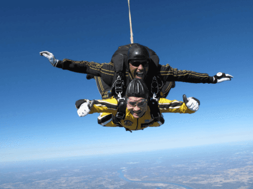 A man, giving a thumbs-up sign, is tandem skydiving with an instructor against a clear blue sky.