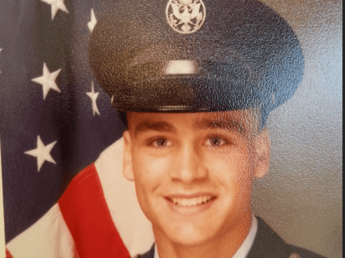 Close-up portrait of a young man in a dark U.S. Air Force dress uniform with a formal cap, smiling against a backdrop of a partially visible American flag.