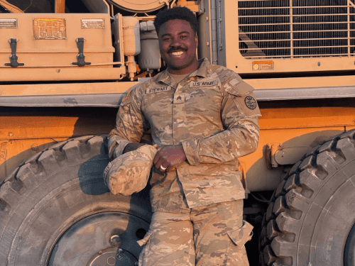 An Army soldier, smiling while leaning on the large tire of a heavy military vehicle, wearing camouflage uniform.