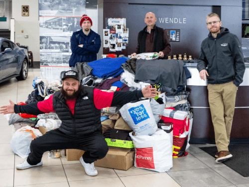 Four men standing inside a Wendle dealership showroom behind a large, accumulated pile of clothing and other donated items, featuring one man crouched in front with his arms wide open.