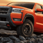 Nissan Frontier PRO-4X pickup truck in orange climbing over jagged rocks on an off-road trail at sunset.
