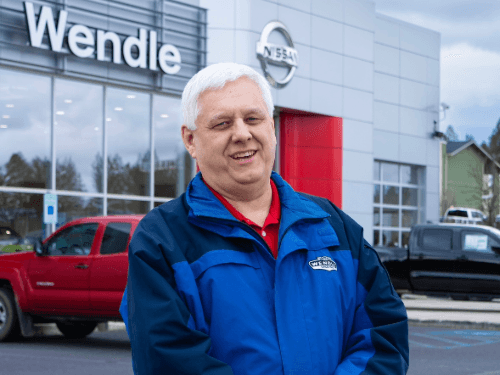 Smiling man in a blue Wendle jacket stands before a Wendle Nissan dealership with cars blurred in the background.