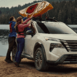 Two people loading a yellow kayak onto the roof rack of a white Nissan Rogue parked on a sandy shore by a scenic lake.