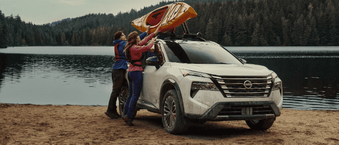 Two people loading a yellow kayak onto the roof rack of a white Nissan Rogue parked on a sandy shore by a scenic lake.
