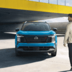 A man walks toward a blue Nissan Kicks parked under a modern yellow bridge structure in a bright, open concrete space.