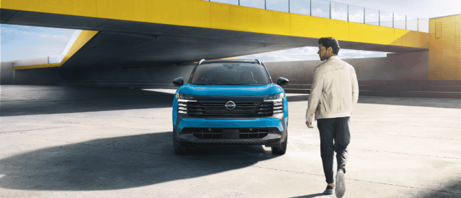 A man walks toward a blue Nissan Kicks parked under a modern yellow bridge structure in a bright, open concrete space.