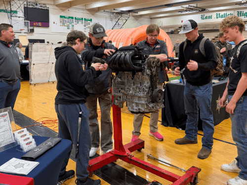 A group of students observing a car engine mounted on a stand at a career fair in a gymnasium.