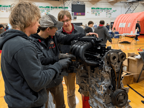 Three students using an impact driver to work on a car engine display during a trades night event in a gymnasium.