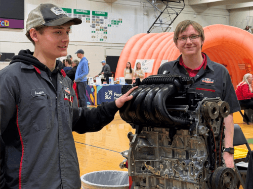 Two students wearing shop shirts standing next to a car engine display on a stand during a trades night career fair.