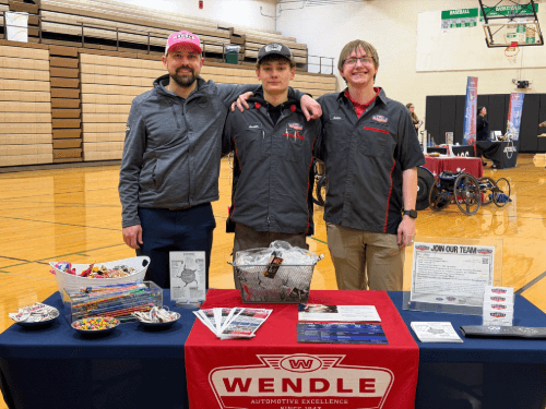 Three men posing together behind a Wendle recruitment table with brochures and candy at a high school career fair.