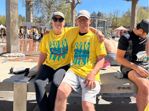 A smiling couple wearing matching yellow 2026 Bloomsday finisher t-shirts sitting together on a bench outdoors.
