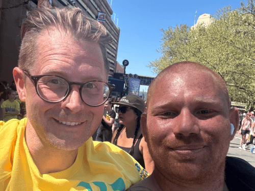 A close-up selfie of two smiling men posing together outdoors on a crowded city street during the Bloomsday run.