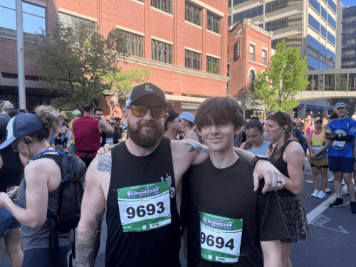 A smiling man and a youth posing together with race bibs on a crowded downtown street for the Bloomsday run.