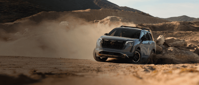 A grey Nissan Pathfinder Rock Creek kicking up dust while driving off-road over a rugged, rocky desert terrain.
