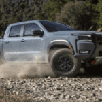 Grey Nissan Frontier PRO-4X pickup truck kicks up dust while speeding along a rugged, rocky off-road trail with green trees.