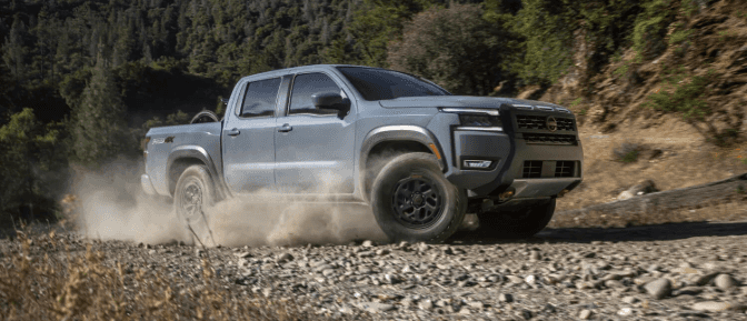 Grey Nissan Frontier PRO-4X pickup truck kicks up dust while speeding along a rugged, rocky off-road trail with green trees.