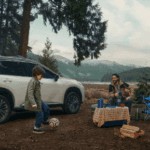 White Nissan Rogue SUV parked at a mountain campsite with a family, blue tent and a child playing soccer near a lake.