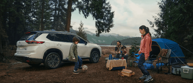 White Nissan Rogue SUV parked at a mountain campsite with a family, blue tent and a child playing soccer near a lake.