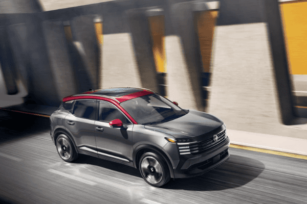 Two-tone Nissan Kicks SUV with a grey body and red roof, driving along a city street lined with modern architecture.
