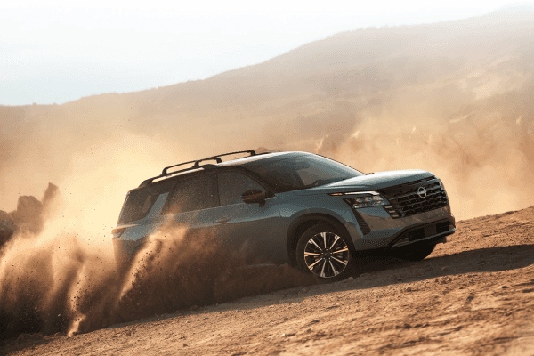 A grey Nissan Pathfinder SUV driving on a dirt terrain, kicking up a cloud of dust under warm, hazy sunlight with hills in the background.