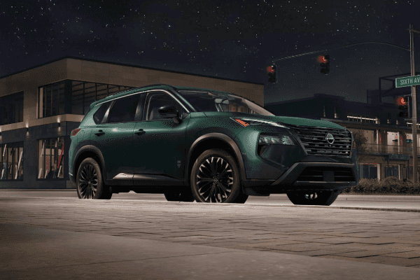 Dark green Nissan Rogue SUV parked along a city street curb at night in front of modern buildings.