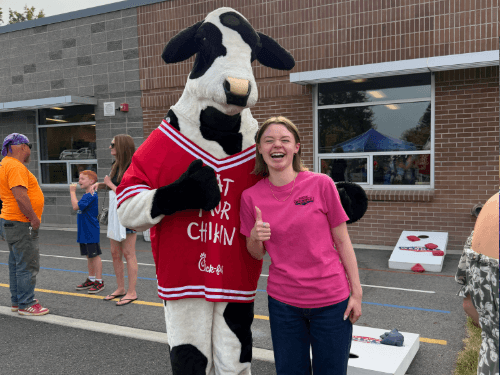 A smiling person in a pink shirt giving a thumbs-up next to a cow mascot wearing a red jersey at an outdoor event.