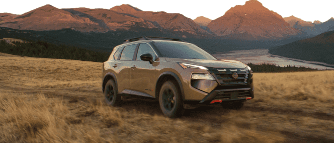 Nissan Rogue Rock Creek driving on an off-road trail through a field with rugged mountain peaks in the background.