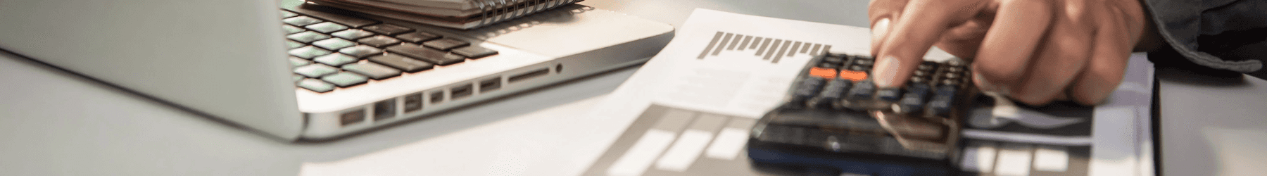 Close-up of a person using a calculator next to financial documents and a laptop computer on a desk.