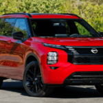 Red 2026 Nissan Rogue Plug-In Hybrid SUV parked outdoors with green foliage in the background.