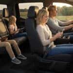 Blonde family seated inside a pickup truck interior at sunset, parents in front wearing seat belts while children in back use tablets, rural Wisconsin farmland visible through the windows.