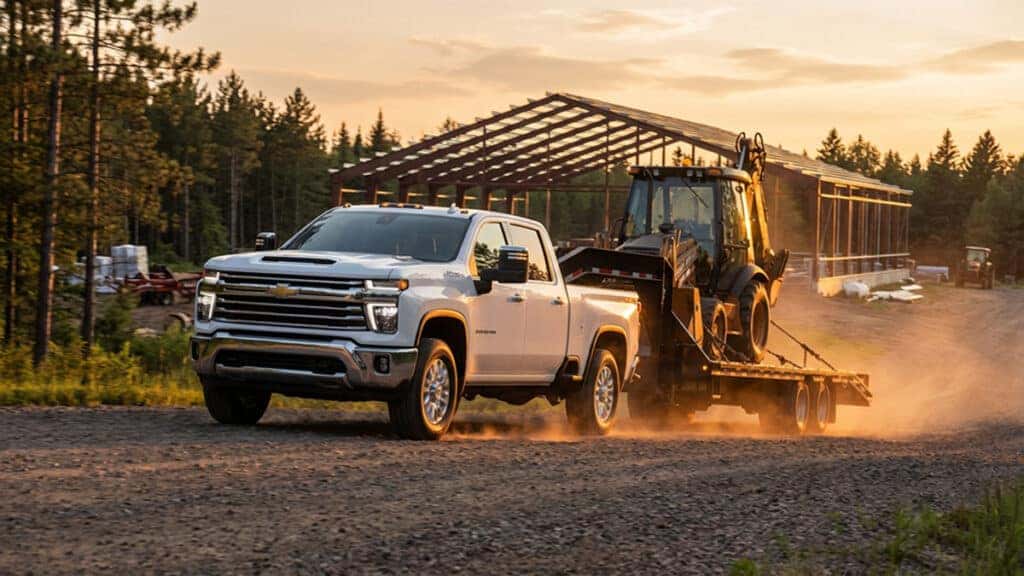 Chevrolet Silverado 2500 HD ripping up a gravel road at golden hour in Northern Wisconsin, towing a fifth wheel trailer hauling a secured backhoe with a free stall dairy barn under construction in the background.