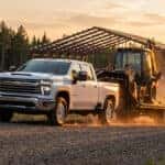 Chevrolet Silverado 2500 HD ripping up a gravel road at golden hour in Northern Wisconsin, towing a fifth wheel trailer hauling a secured backhoe with a free stall dairy barn under construction in the background.